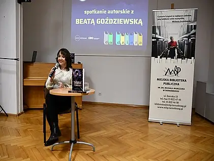 Beata Goździewska siedzi na krześli i mówi do uczestników spotkania